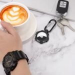 Husa de protectie neagra pentru AirTag, o mana cu un ceas si o ceasca de cafea